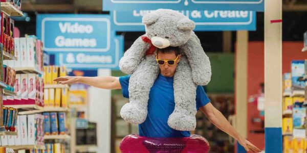 A man in a blue t-shirt, wearing a pink inflatable around his waist, a pair of yellow glasses, and a large grey stuffed bear on his shoulders
