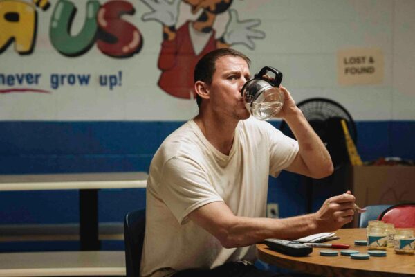A man in a white t-shirt sits at a table in front of a partially obscured Toys "R" Us mural, drinking water from a coffee carafe