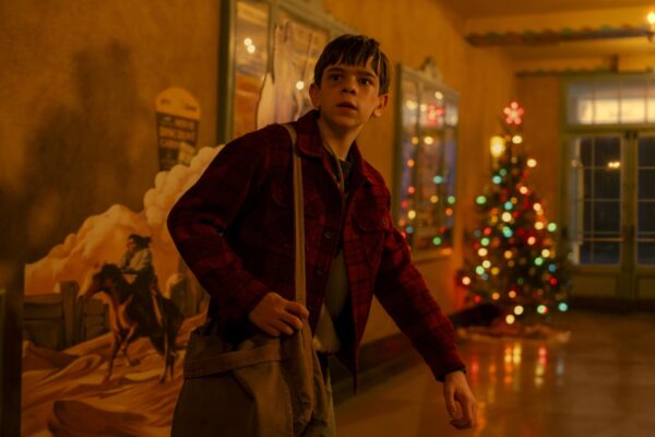 A disheveled pre-teen boy carrying a bag stands in a hallway with a Christmas tree in the background