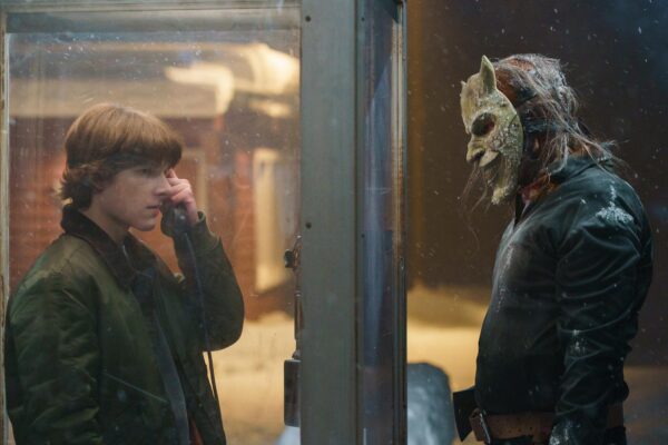 A teen boy (l) with shaggy hair holds a pay phone to his ear as a masked figure with long hair watches on the outside (r)
