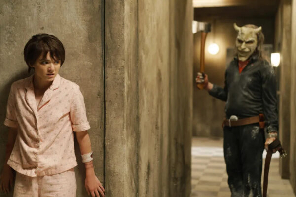 A pre-teen girl in pink pajamas hides behind a wall as a masked figure holding a raised axe approaches