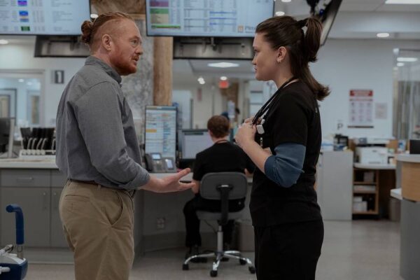 A man with red hair in a bun shakes the hand of a young woman in blue scrubs with her dark hair in a ponytail