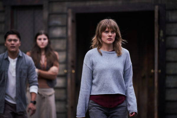 A dirty blonde woman in a grey/lavender long sleeve shirt stands in front of an Asian man and a dark haired woman with her arms crossed in the background