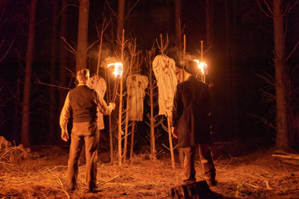 At night, in a scene lit by torches, two men stand in front of tree branches stuck in the ground, adorned with bloody dresses on them