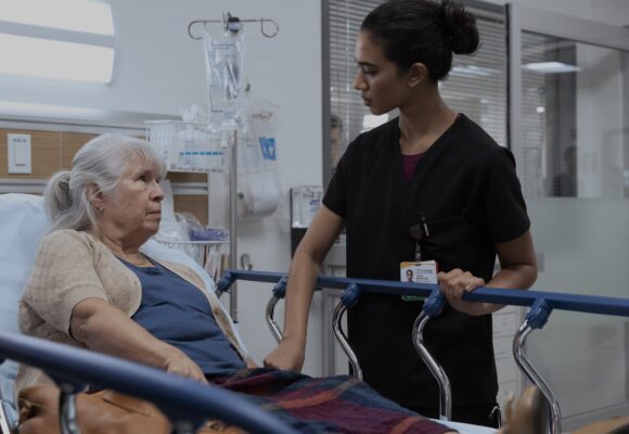 A dark skinned doctor in black scrubs reassures an elderly white woman sitting upright in a medical bed