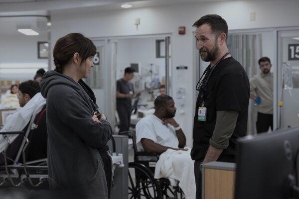 A woman wearing a grey hoodie with her dark haired tied in a ponytail stands with her arms crossed in front of a male doctor in black scrubs