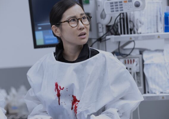 An asian woman wearing glasses and a white surgical gown with two spots of blood on it
