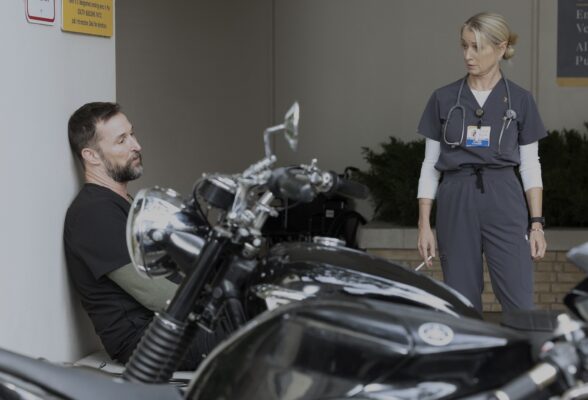 A male doctor with greying facial hair (L) sits behind a motorcycle as a blonde nurse stands (r)