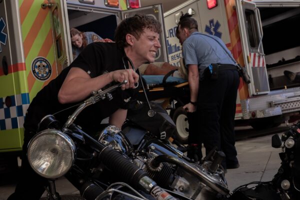 A young white man in black medical scrubs struggles to lift a motorcycle that's fallen over