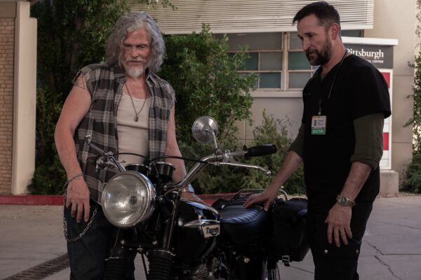 A gray haired man in a white tank top and cut off plaid shirt surveys a motorcycle next to a dark haired doctor with a beard dressed in black scrubs