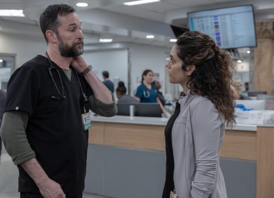 A woman with curly dark brown hair wearing a off-white sweater over black scrubs looks at a taller man with a trimmed beard in black scrubs