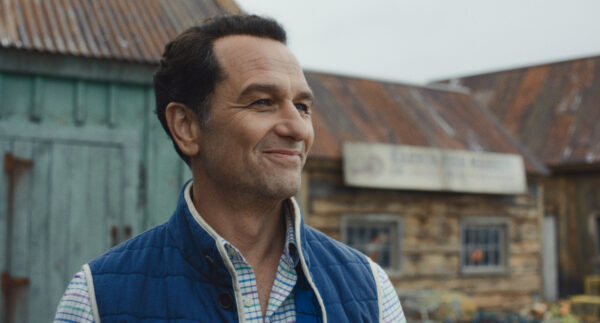 A dark haired man in a blue vest and blue button down shirt gives a forced smile