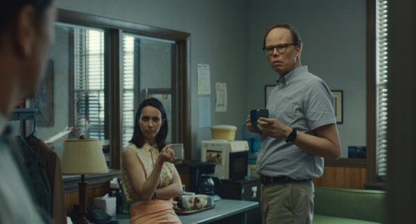 A pair of white co-workers - a woman with dark hair (L) and a tall man with glasses (R) hold coffee cups