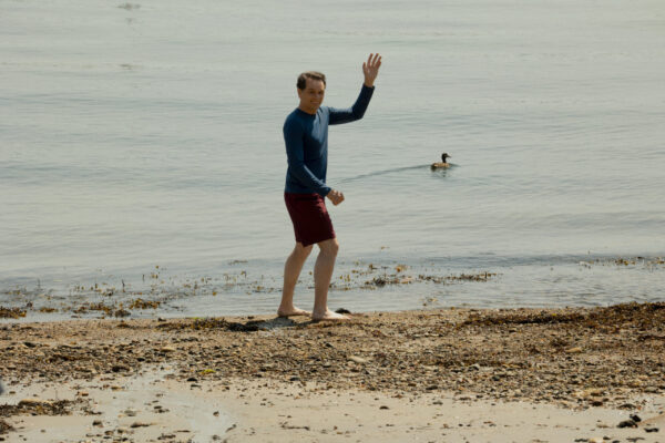 A dark haired man in a blue wet suit, waving on the beach