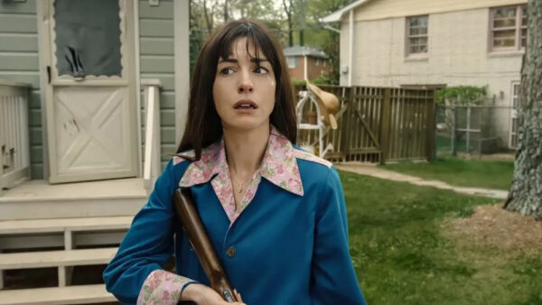 A dark haired woman in a blue sweater pauses cautiously on the lawn of a suburban house