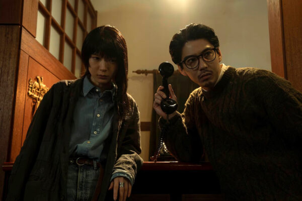 A Japanese woman with asymmetrical haircut crouches next to a Japanese man wearing dark rimmed glasses in a hallway