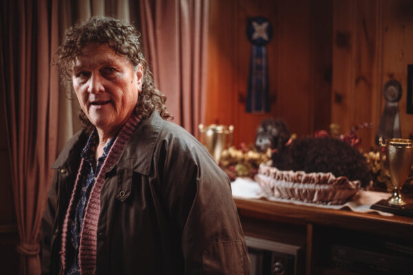 A middle aged woman with shoulder length brown hair wearing a leather jacket, standing in a red room