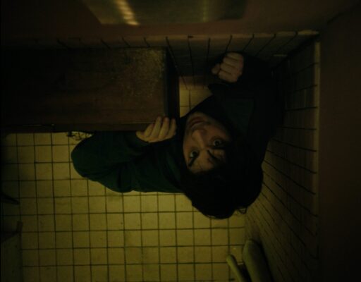 An overhead shot of a dark haired young man reaching up towards a vent in the ceiling