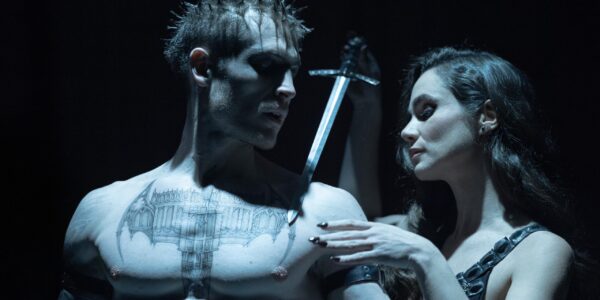 A medium shot of a man from the pecs up as he looks to his right at a dark haired woman in a tank top pressing a knife against his tattoo'd chest