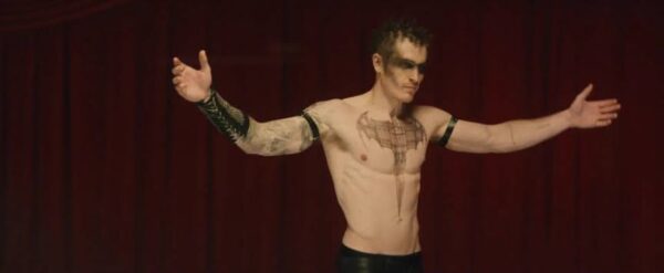 A shirtless, tattoo'd man in black leather pants, arms outstretched, with black eye makeup, standing in front of a red curtain