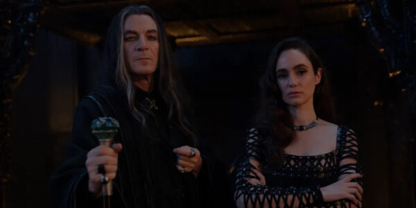 A man with long black and grey hair stands next to a woman in a low cut lace dress