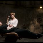 A woman in a white shirt examines a man in black on a reclining chair