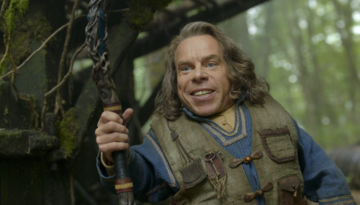 Warwick Davis as Willow, holding a walking stick in the forest