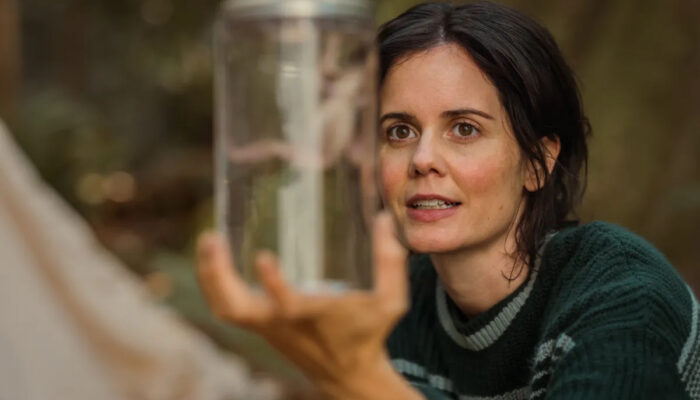 A brunette woman holds a glass jar containing a frog