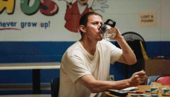 A man in a white t-shirt sits at a table in front of a partially obscured Toys "R" Us mural, drinking water from a coffee carafe