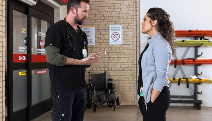A tall dark haired man with a beard in blue scrubs faces a woman with curly dark hair in a light grey shirt and black pants in front of the ER doors