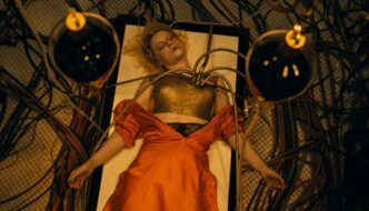 A dead woman with blonde hair, wearing an orange dress pulled down to reveal a gold tanktop, with wires plugged into her chair