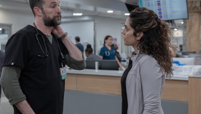 A woman with curly dark brown hair wearing a off-white sweater over black scrubs looks at a taller man with a trimmed beard in black scrubs