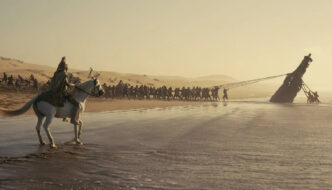 A man in armour sitting on a white horse in the foreground as a line of men raise an enormous wooden horse from the water in the background