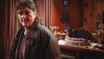 A middle aged woman with shoulder length brown hair wearing a leather jacket, standing in a red room
